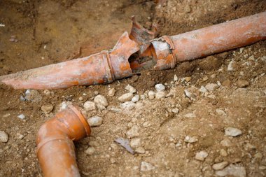 Görüntü, hasarlı turuncu plastik boruların kısmen toprak ve çakıllara gömüldüğünü gösteriyor. Bir boru ciddi şekilde çatlamış ve kırılmış, içinde enkaz var..
