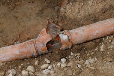Hasarlı bir turuncu boru, kısmen toprak ve çakıllara gömülmüş, iç kısımda ciddi bir çatlak gösteriyor..