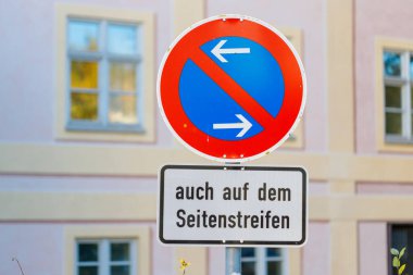 Her iki yönde durmayı yasaklayan dairesel bir tabela, pencereleri olan bir binaya dayanan dikdörtgen bir Alman yazısının üzerinde sergilenmektedir..