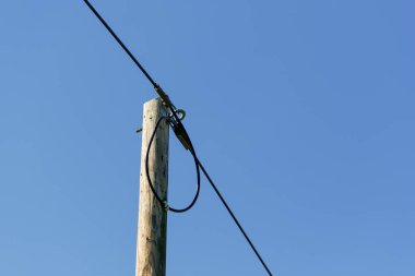 Tahta bir elektrik direği açık mavi gökyüzüne karşı siyah bir kabloyu destekler. Kablonun bir kısmı direğe bağlı..
