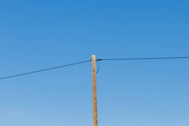 Tek bir ahşap elektrik direği açık mavi gökyüzüne karşı duruyor. Çerçeve boyunca yatay olarak uzanan iki koyu kabloyu destekliyor..