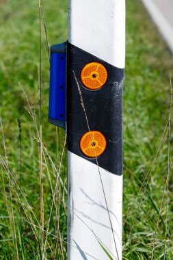 İki turuncu yansıtıcı ve yeşil çimenlerin arka planında mavi yansıtıcı paneli bulunan beyaz ve siyah bölümlü bir yol kenarı işaret direği..