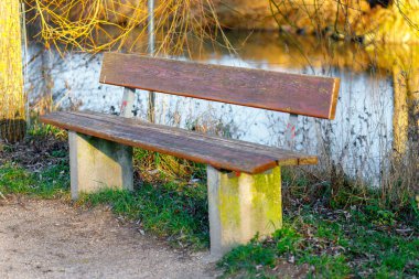 Sonbahar yeşilliklerinde sıcak güneş ışığı olan yansıtıcı bir göletin yanındaki boş ahşap bank