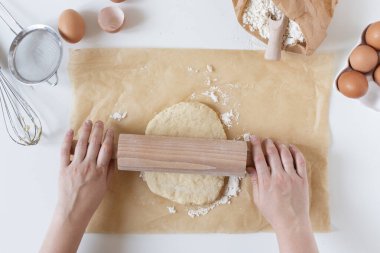 Beyaz masa manzaralı parşömen üzerinde merdaneyle hamuru yuvarlayan kadın elleri. Hamur pişirme için hazırlanıyor.