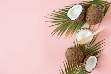 Jar of coconut oil and fresh coconuts with palm leaves on pink background top view with copy space.