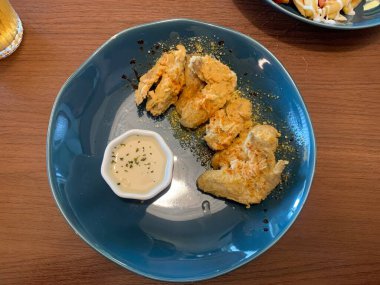 Ahşap masada mavi tabakta servis edilen kızarmış tavuk kanatları. Yanında da otlarla süslenmiş küçük bir tabak kremalı beyaz sos..