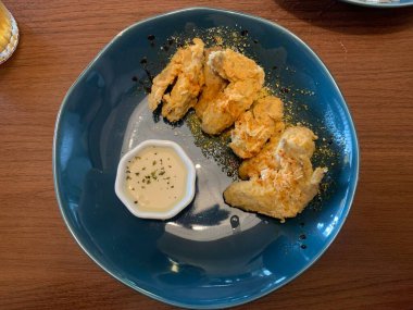 Ahşap masada mavi tabakta servis edilen kızarmış tavuk kanatları. Yanında da otlarla süslenmiş küçük bir tabak kremalı beyaz sos..