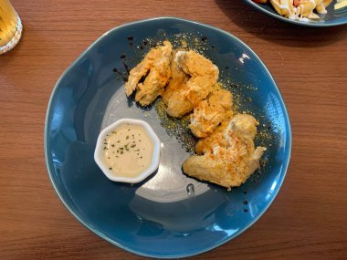 Ahşap masada mavi tabakta servis edilen kızarmış tavuk kanatları. Yanında da otlarla süslenmiş küçük bir tabak kremalı beyaz sos..