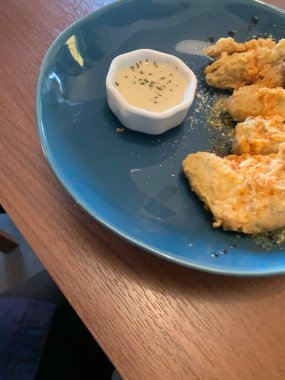 Ahşap masada mavi tabakta servis edilen kızarmış tavuk kanatları. Yanında da otlarla süslenmiş küçük bir tabak kremalı beyaz sos..