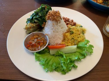 Bebek Betutu rostosu ördeği eşliğinde sunulan beyaz tabakta servis edilen bale yemeği ve üzerine kızarmış arpacık soğanı ile bol miktarda pirinç. Sotelenmiş sebzeler rendelenmiş hindistan cevizi, kızarmış fıstık, salata, marul, salatalık, domates dilimi içerir..