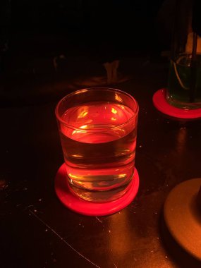 A clear glass contains orange liquid likely water on pink coaster. It is illuminated by warm lighting creating reflective glow.