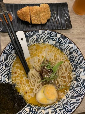 Dilimlenmiş yeşil soğan ve mantarla kaplı erişte ve katsuyla dolu dekoratif bir kase. Et suyu bol kremalı, görünür yağ damlacıkları gibi görünüyor.