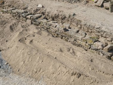 Kayaların düzenli olarak kumlu toprağa istiflendiği, geleneksel yapı yöntemleri, istikrar ve mirasın korunmasını gösteren rekonstrüksiyon altındaki antik taş duvar..