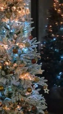 Işıkları ve altın süslemeleri olan süslü bir Noel ağacı bir pencereye yansıyan kapalı bir alanda duruyor. Sahne, gece vakti özel bir evde festival detaylarını ve tatil ruhunu yakalıyor..