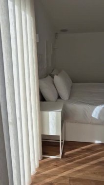 Yumuşak gün ışığı filtreleri perdelerin arasından rahat bir yatak odasına, düzgün yapılmış çift kişilik yatak, beyaz çarşaflar ve yastıklar, modern bir evde dinlenmek ve dinlenmek için sakin, davetkar bir yer yaratıyor..