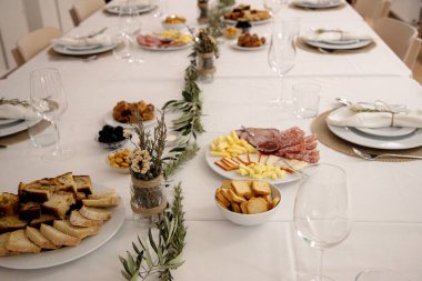 Beyaz tabaklar, şarap bardakları ve kırsal yeşilliklerle dolu şenlikli bir yemek masası hazırlanırken, çeşitli peynir, kurutulmuş et, ekmek, zeytin ve abur cuburlar aileleri ve arkadaşlarını kutlama yemeği için bir araya getirmeye davet ediyor..