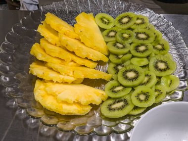 Profesyonel bir mutfakta cam bir tabağa yerleştirilmiş taze ananas ve kivi dilimleri sağlıklı atıştırmalık olarak hazırlanmış canlı tropikal meyve tabağı veya restoran servisi için tatlı.