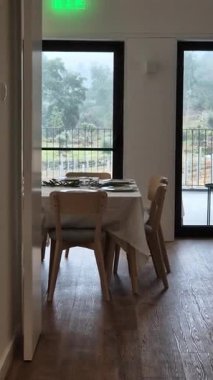 Geniş pencereli, yağmurlu kırlara bakan, sakin bir kırsal kesimde misafirleri ağırlamaya hazır yemek masası ile rahat bir yemek odası..