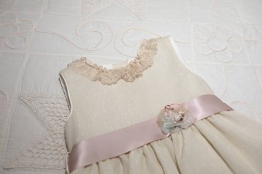 Küçük bir kızın tören elbisesi, dantel yaka, saten pembe kemer ve çiçek aplikasyonu, özel bir durum için özel işlemeli beyaz bir yatak örtüsü üzerinde hazır..