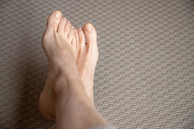 Çıplak gerçek kadın ayakları, evdeki desenli bej halıda ayakta ve çaprazlanmış, temiz tırnaklar ve yumuşak ışıkta vurgulanmış doğal ten rengi, sağlık ve yaşam tarzı kavramları için basit vücut ayrıntıları..