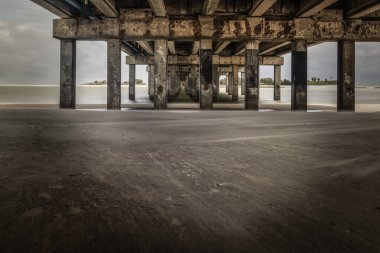 Atlantik kıyısındaki bir Florida rıhtımının altında sakin ve uzun pozlu bir manzara. Bileşim, endüstriyel yapıyı doğal sakinlikle harmanlayarak gelgit, doku ve ışığın ritmini yakalıyor..