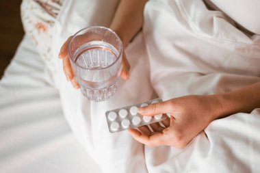 Yatakta bir bardak su ve beyaz yatak ve yumuşak ışıklandırmayla çevrili bir paket tablet tutan bir kadın bir an dinlenme ve bakım öneriyor.