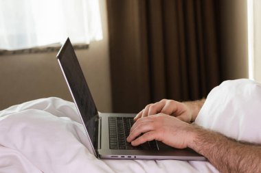 Beyaz çarşaflar ve kahverengi perdelerle çevrili yatağında dinlenirken dizüstü bilgisayarda yazı yazan kişi sahneyi aydınlatıyor.