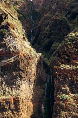 Şelale, dağlık bir bölgedeki canlı yeşil bitki örtüsüyle çevrili dik kayalıklardan aşağı akar ve doğal manzarayı ve jeolojik özellikleri vurgular.