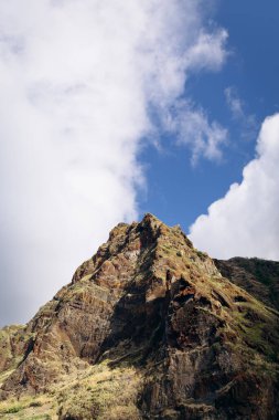 Dağ zirvesinde engebeli uçurumlar ve parlak mavi gökyüzüne karşı yemyeşil alanlar, bölgenin doğal manzarasını ve jeolojik özelliklerini vurguluyor.