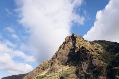Dağ zirvesinde engebeli uçurumlar ve canlı yeşillikler kabarık bulutlu berrak mavi gökyüzüne karşı çarpıcı bir tropikal manzara sergiliyor.