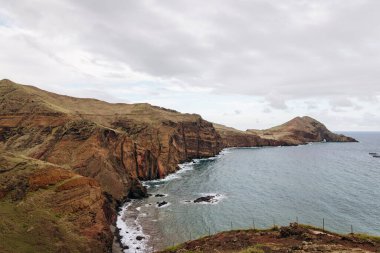 Dik uçurumların okyanusla buluştuğu kıyı manzarası, bulutlu bir gökyüzü altında toprak tonlarının ve nazik dalgaların karışımını sergiliyor.
