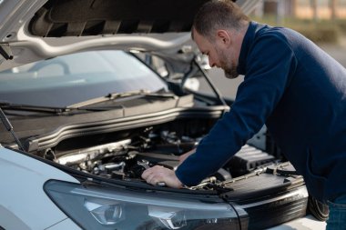Erkek tamirci kaputun altındaki motor parçalarını inceliyor, mavi bir ceket giyiyor, açık havada doğal ışıklandırmalı bir ortamda.