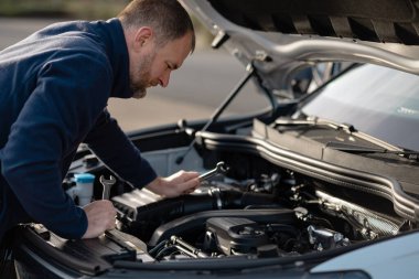 Adam, motor parçalarını elinde aletlerle, kaputu açık bir ortamda doğal ışık ve bulanık bir arka planla inceliyor.