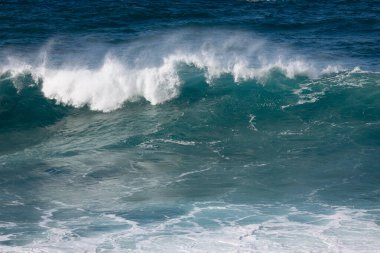 Kıyı şeridindeki büyük dalgalar beyaz köpük ve su sıçratır. Derin mavi su ve arka planda berrak bir gökyüzü vardır.