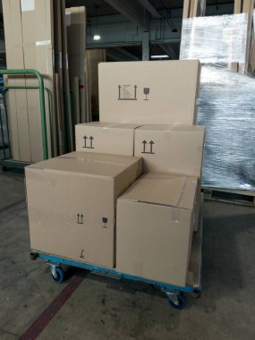 Karton kutular, arka planda ahşap paletler ve depolama malzemeleriyle çevrili bir depodaki mavi bir dolinin üzerine istiflenir.