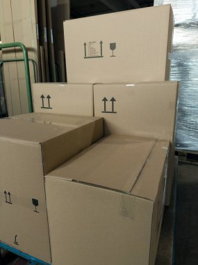 Depoda görülebilir kullanım sembolleri olan, farklı boyutlarda ve yönlere sahip, yanında palet ve plastik ambalaj bulunan karton kutular.