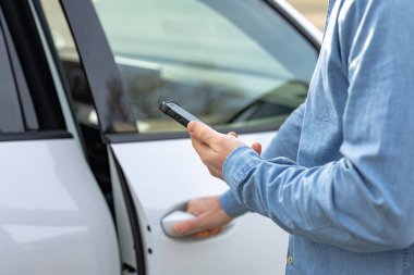 Akıllı Şehir Taşımacılığı. Mavi tişörtlü bir adam, bulanık bir çevrede beyaz bir arabanın kapısını açarken akıllı telefon tutuyor.