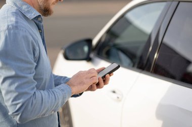 Mobil araba kiralama servisi. Mavi tişörtlü bir adam, şehir ortamında beyaz bir arabanın yanında akıllı bir telefonla, mobil araba kiralama hizmeti bağlamında iletişim kuruyor.