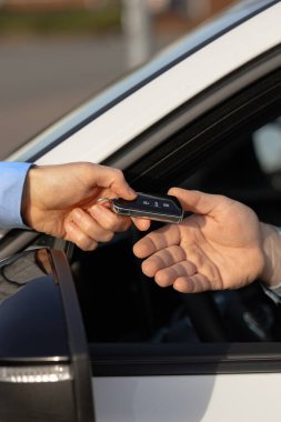 İki el araba kiralama anahtarlarını bir arabanın camında değiş tokuş ederken, arka planda modern bir araba ve açık hava ortamını ön plana çıkarıyor.