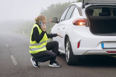 Her gün stres. Yansıtıcı güvenlik yeleği giyen kadın yol kenarında beyaz bir arabanın yanında diz çöküp sisli bir ortamda telefonla konuşuyor.