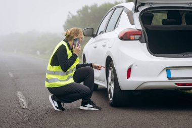 Yol kenarı desteği. Yansıtıcı yelek giyen bir kadın yol kenarında beyaz bir arabanın yanında diz çöküp sisli bir şekilde cep telefonu kullanırken lastiği inceliyor.