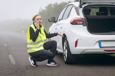 Her gün stres. Yansıtıcı yelek giyen kadın yol kenarında beyaz arabanın yanına diz çöker, telefonda konuşur ve arka planda sisli bir manzara vardır.