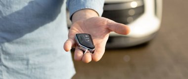 Bireyin elinde, parlak güneş ışığı altında arka planda biraz bulanık odaklı beyaz bir araba bulunan bir araba anahtarı bulunuyor. Pankart