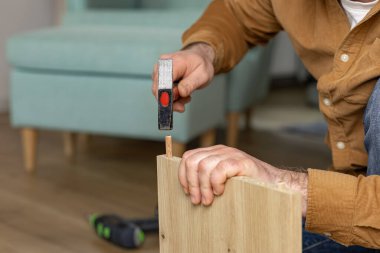 Erkek marangoz, modern bir oturma odasında, yerde açık mavi bir koltuk ve aletlerle ahşap mobilyalar monte etmek için çekiç kullanıyor.