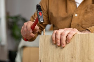 Bir adam dolap mobilyası monte ediyor. Kahverengi gömlekli bir adam çekiçle mobilya monte ediyor, rahat bir ev dekorunda ahşap parçaları güvence altına alıyor.