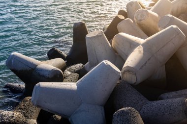 Kayalık bir kıyı şeridinde istiflenmiş beton tetrapodlar, kısmen suya batmış, güneş ışığı yüzeyden yansıyarak bir kıyı ortamı yaratıyor.