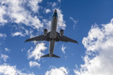 Aşağıdan görülen mavi gökyüzü ve etrafını saran bulutlarla uçağın kanatları ve motorları net bir şekilde vurgulanıyor.