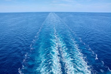 View of the wake of a large ship moving away in the blue ocean. The waves and white foam create a trail extending to the distant horizon.