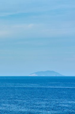 Vertical seascape with a deep blue sea and clear sky. A distant island on the horizon suggests solitude, escape, and the beauty of nature.