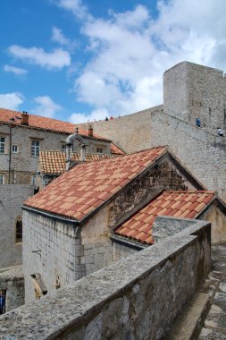 Hırvatistan 'ın Dubrovnik kentinde kentin duvar yürüyüşü manzarası. Sergide ortaçağ taş mimarisi, kırmızı kiremitli çatılar ve mavi bir gökyüzünün altındaki kale yer alıyor..